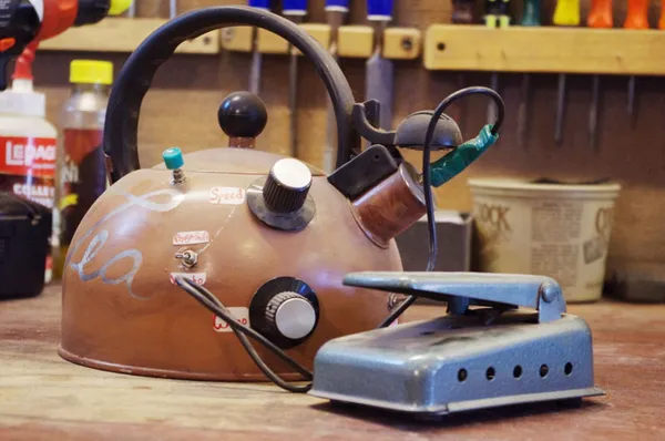 A rust-colored teapot on a workbench with guitar electronics and a foot pedal added to it to give the impression that it could generate sound.