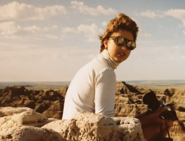 A young woman wearing glacier glasses sitting on a rock in the Badlands with a blue sky and horizon spread out behind her.