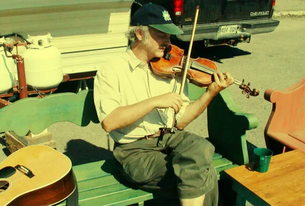 Middle-aged fiddler with baseball cap playing on a green wooden bench in a parking lot.