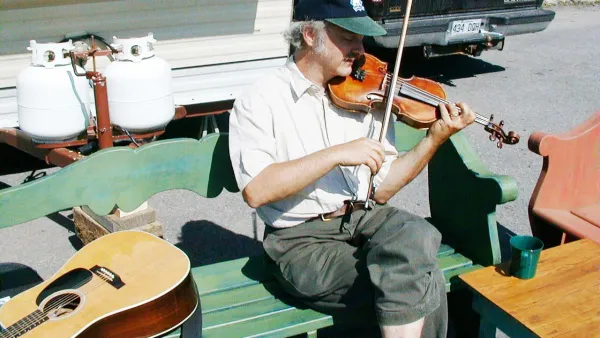 Fiddler wearing baseball cap, playing on a bench in front of a camper