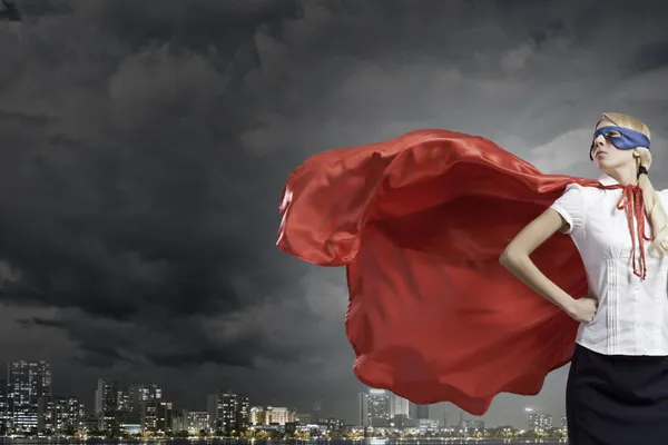 A woman wearing a red super-hero cape and blue eye mask in front of a gray, cloudy cityscape