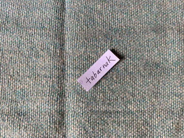 The French swear word "tabarnak" written in black on a piece of pink paper, laying on a background of handwoven, wool cloth.